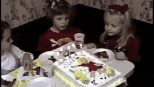 Ein GIF von einem Heimvideo einer Geburtstagsfeier aus den 1970ern oder 1980ern mit drei weißen Kindern im Alter von acht oder neun Jahren, die an einem Tisch sitzen. Zwei der Kinder blasen die Kerzen auf einem riesigen rechteckigen Geburtstagskuchen aus. Von den ausgeblasenen Kerzen steigt Rauch auf, den ein drittes Kind mit der Hand wegwedelt.